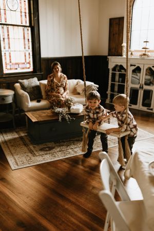two young children play on an indoor swing as their mother lovingly looks on from the couch as light floods through the window