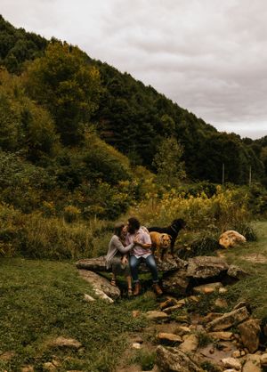 a couple on a rock over a creek with the mountains and flowers in the background as their dog keeps watch in Western North Carolina