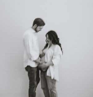 a black and white photo of a mother and father to be in front of a white background while they place their hands on the growing belly