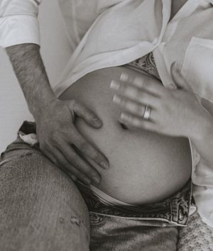 a black and white photo of a woman's pregnant belly in an unbuttoned white shirt both her hand and a hand of a man are placed on her stomach