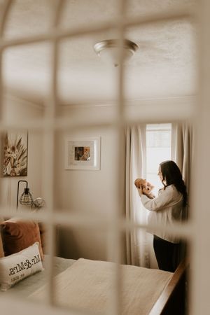 as seen through a mirror on the wall a mother stands in front of the window and looks down at her infant child