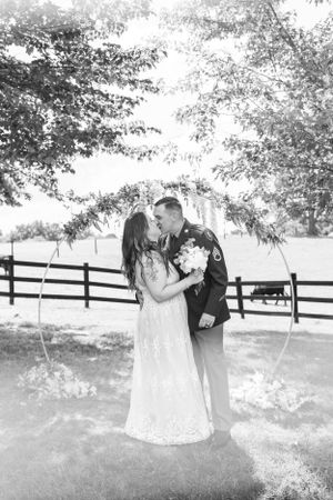 an army man kisses his bride in black and white in front of a flower arch outside by a fence and field