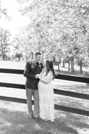 an army man and his bride lean against a fence by a pasture in black and white