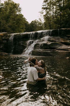 a couple embrace and kiss in the waters below a waterfall