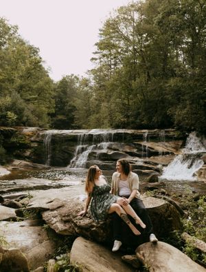 a couple sits on a large rock and looks at each other, a waterfall can be seen in the background