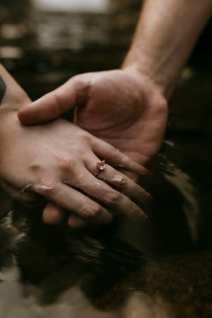 a couple holds hands half in the water with an engagement ring on the top hand