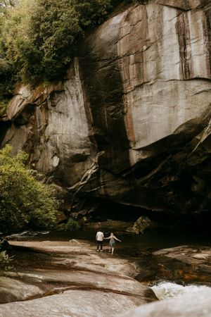 a couple walks in the creek below a giant rock face in the mountains of Asheville