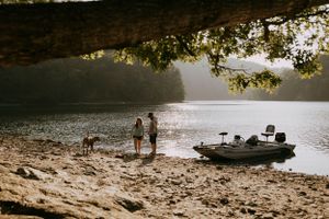 a family stands on a rocky beach at the lake their boat can be seen in the water and a large tree frames the top of the photo
