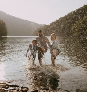a family of three kick and splash water up in the air as they stand in the lake that is surrounded by mountains