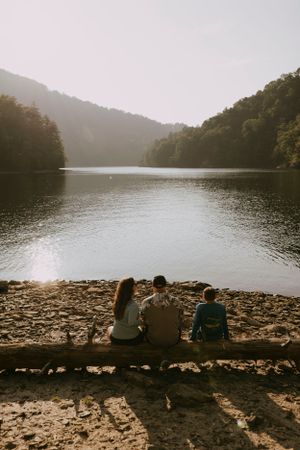a family of three sit on a log on a rocky beach overlooking a lake in the mountains