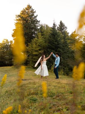 a groom spins his bride in the grass with yellow flowers framing them and her veil flying in the wind in front of the pine trees