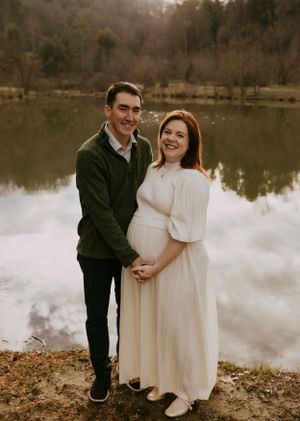 a couple who is expecting a baby stand together cradling her belly in front of a pond
