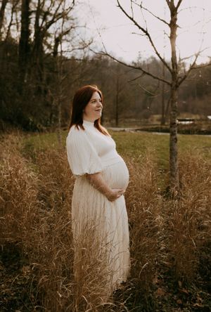 a pregnant woman stand amid the tall grass and looks off into the distance