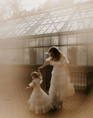 a mother and child spin their flowy white dresses by a greenhouse on a brick path