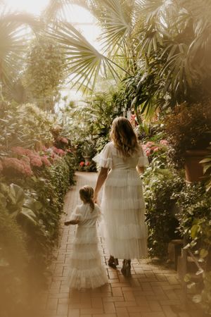 a mother and child in flowy dresses walk through a vivid greenhouse that is cascaded with golden light