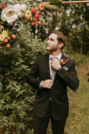 a proud groom fixes his tie under the flower arch