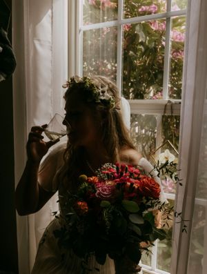a bride sits in a dark room on the windows ledge with her bouquet and a glass of wine