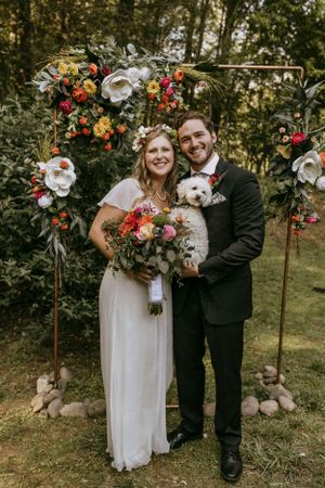 a bride and groom smile proudly holding their dog in front of a flower arch