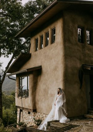 A bride leans against the side of a uniquely crafted house in the mountains