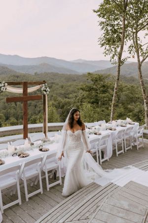 a bride walks along the table that will hold her wedding guests for dinner on a large deck overlooking the Blueridge mountains of Asheville