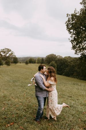a man and woman embrace she has a dress and her leg kicked up as she holds her shoes over his shoulders. they are in a field in Asheville