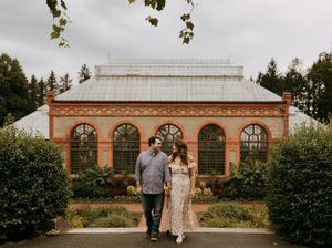 a man and woman stand between two bushes and in front of the conservatory greenhouse at the Biltmore Estate in Asheville North Carolina