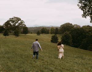 a man and woman walk through a large field full of trees that is in front of the Blueridge Mountains of Western North Carolina in Asheville