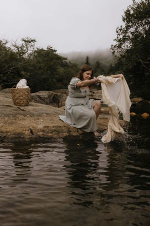 a woman leans down on a big rock with a white sheet she is preparing to wash in the waters below in Western North Carolina near Asheville