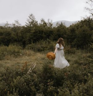 a woman with a dress and a basket walks through a open field surrounded by bushes in Western North Carolina near Asheville