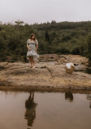 a woman in a dress on a big rock gets ready to lean down to the water to wash her laundry in Western North Carolina near Asheville