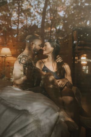 a couple sit on the bed holding each other close as the reflection of the tall trees outside cover them in Western North Carolina Asheville