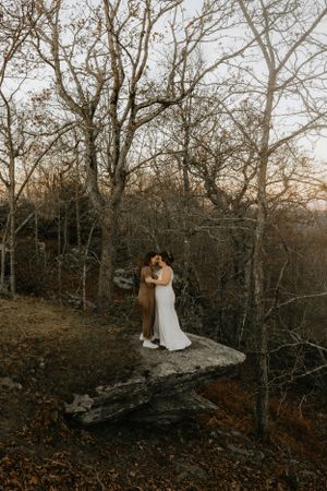 two brides embrace on a rock in the woods