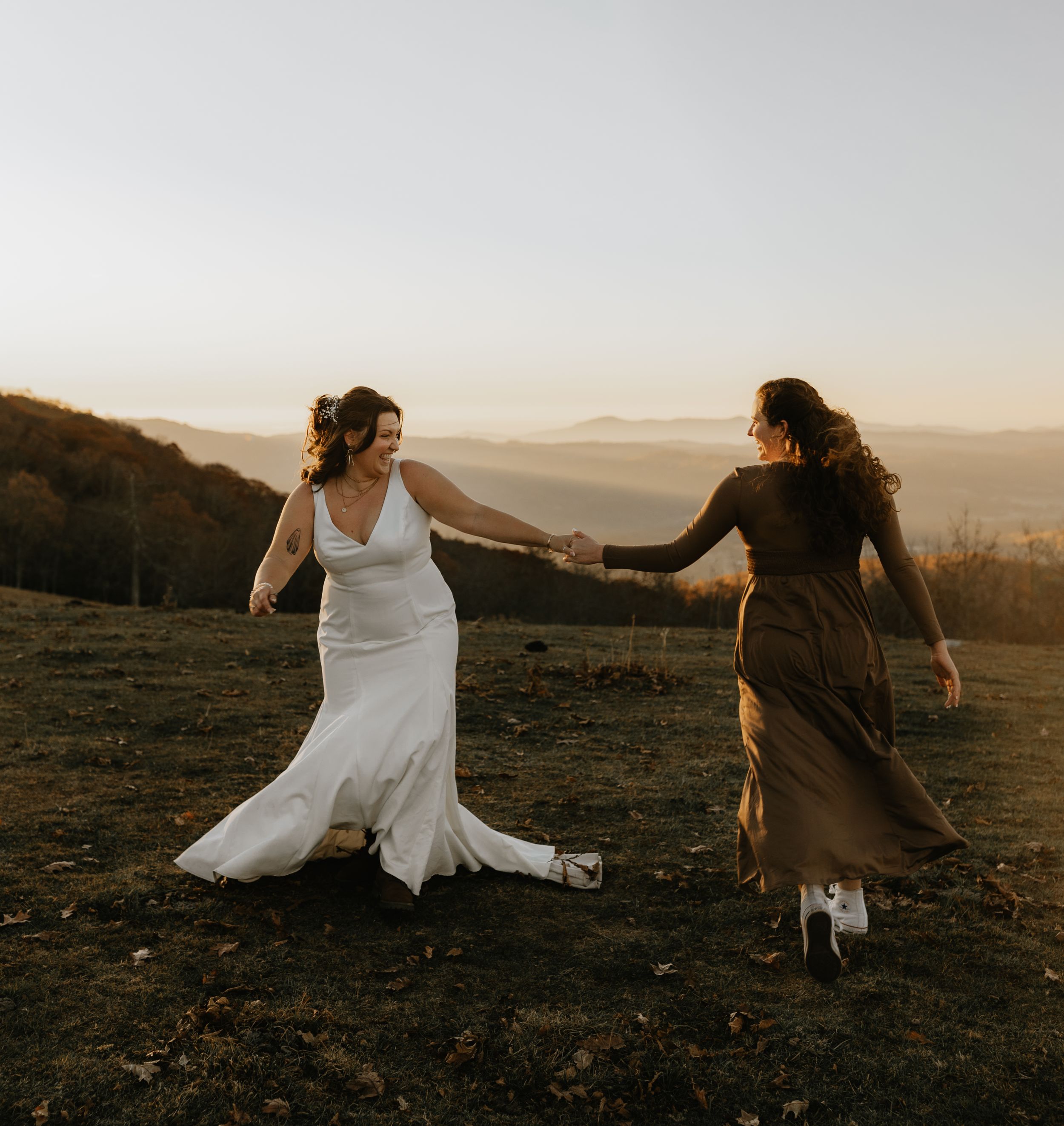 two brides holding hands in the sunrise on top of the mountain in their dresses with the Blueridge mountains in the background