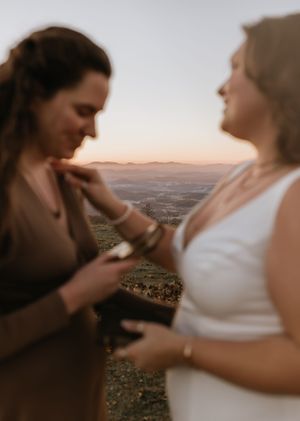 two brides out of focus with the focal point being the landscape of mountains below them in the sunrise