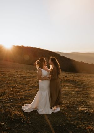 two brides hug in an open field with mountains and the sunrise coming over the ridge