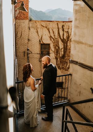 a couple shares a longing look on the stairs of a brick building the blue ridge mountains can be seen in the background