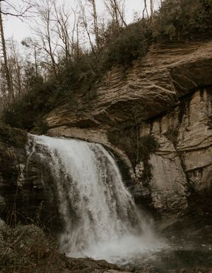 a large roaring waterfall with a large rock wall beside it plummets into the river below in Asheville North Carolina