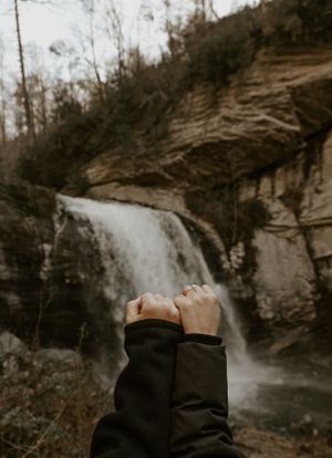a couple hold their hands up in front of a waterfall to celebrate their engagement