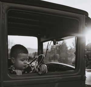 a monochrome shot of a little boy in a vintage truck driving his toy car along the window