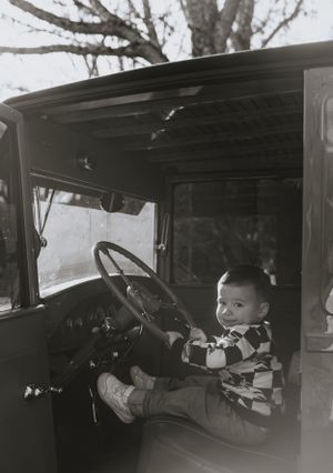 a monochrome shot of a little boy in a checkered sweater reaches the steering wheel of a vintage truck