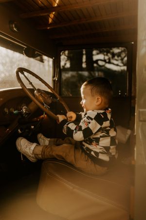 a little boy in a checkered sweater reaches the steering wheel of a vintage truck