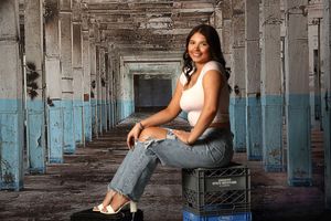 A person sits on a chair in an abandoned industrial hallway with blue-painted walls and concrete pillars.
