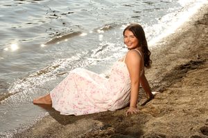 A soft pink floral dress adorns someone posing on a sandy beach beside gentle waves.