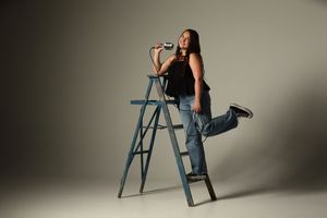 Woman in jeans and black top posing on a blue step ladder against a gray studio background.