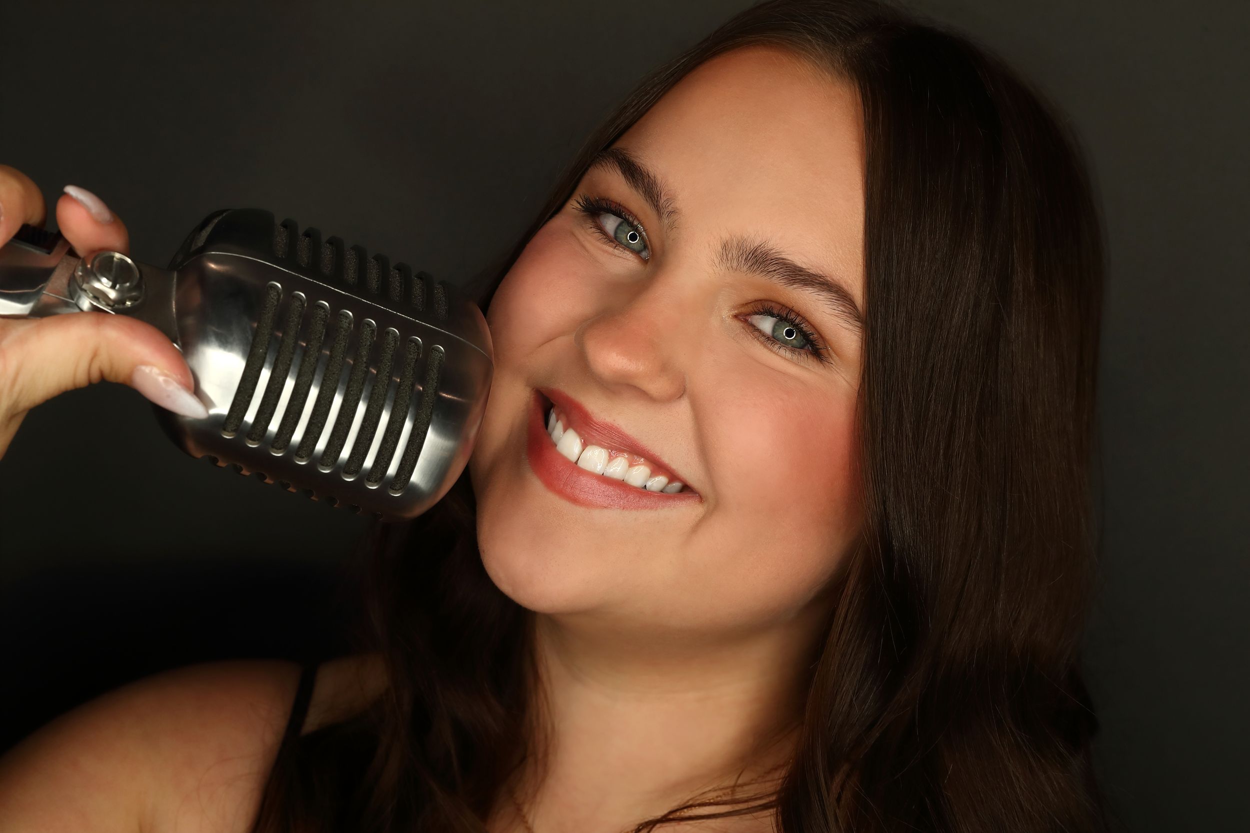Smiling brunette woman holding a vintage silver microphone close to her face against a dark background.
