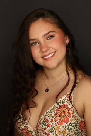 Smiling young woman with long brown hair wearing a floral dress and necklace against a dark background.