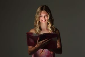 Someone in pink dress smiling while reading from a book in dramatic lighting.