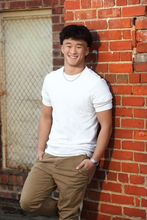 Young Asian man in white t-shirt and khaki pants smiling while leaning against a red brick wall outdoors.
