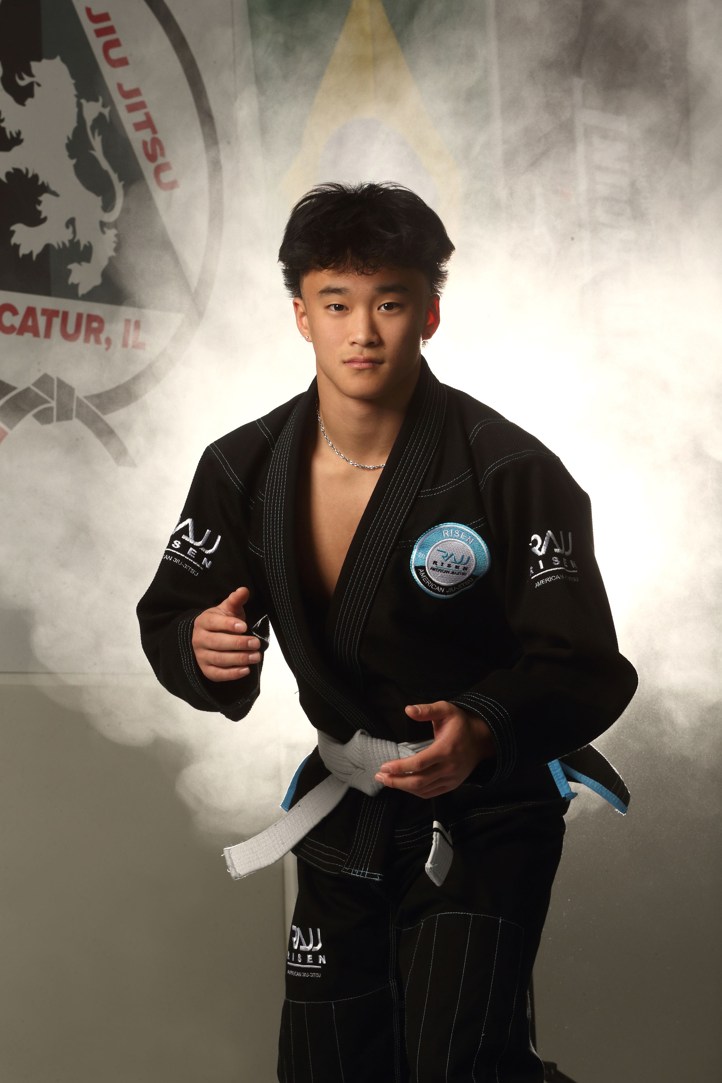 Young martial artist in black BJJ gi with white belt poses confidently in front of Jiu Jitsu academy backdrop.
