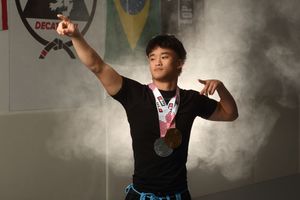 Young male athlete poses proudly wearing multiple medals in a gym with Brazilian flag and smoke effects in background.